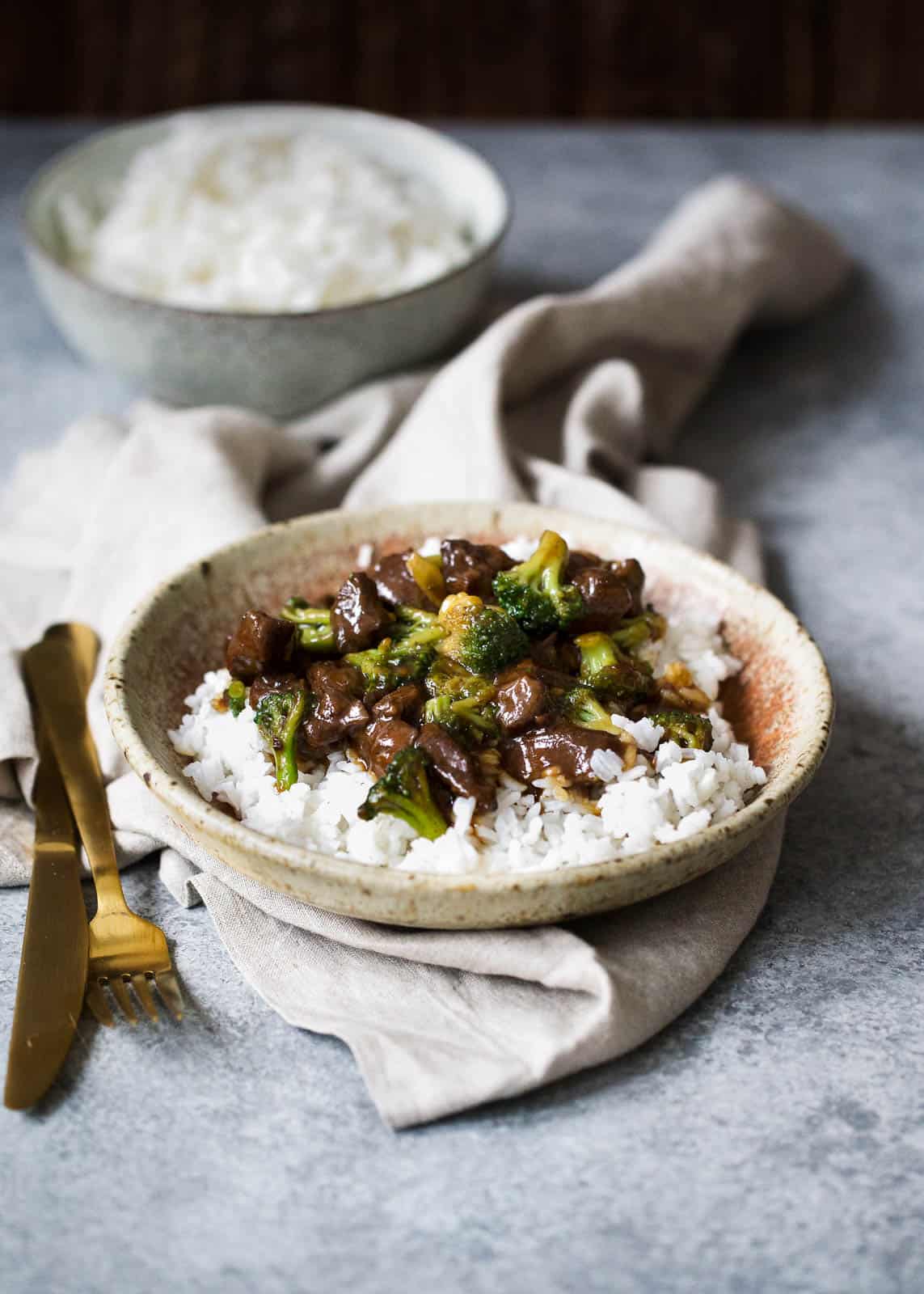 beef with broccoli mixture in a tan speckled bowl with white rice. the bowl sits on top of a beige linen towel with a gold fork and knife on the side and a bowl of white rice in the background.
