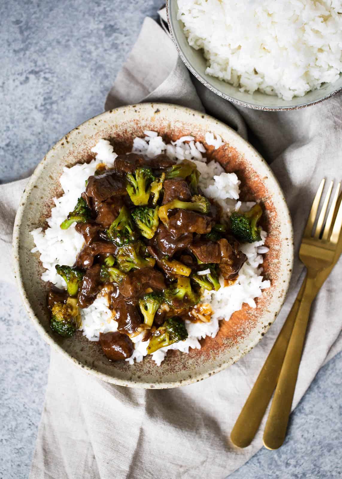 overhead shot of beef and broccoli mixture over rice in a tan speckled bowl with a rust coloring interior. the bowl sits on top of a beige linen towel with a gold fork and knife and bowl of white rice in the upper right corner.