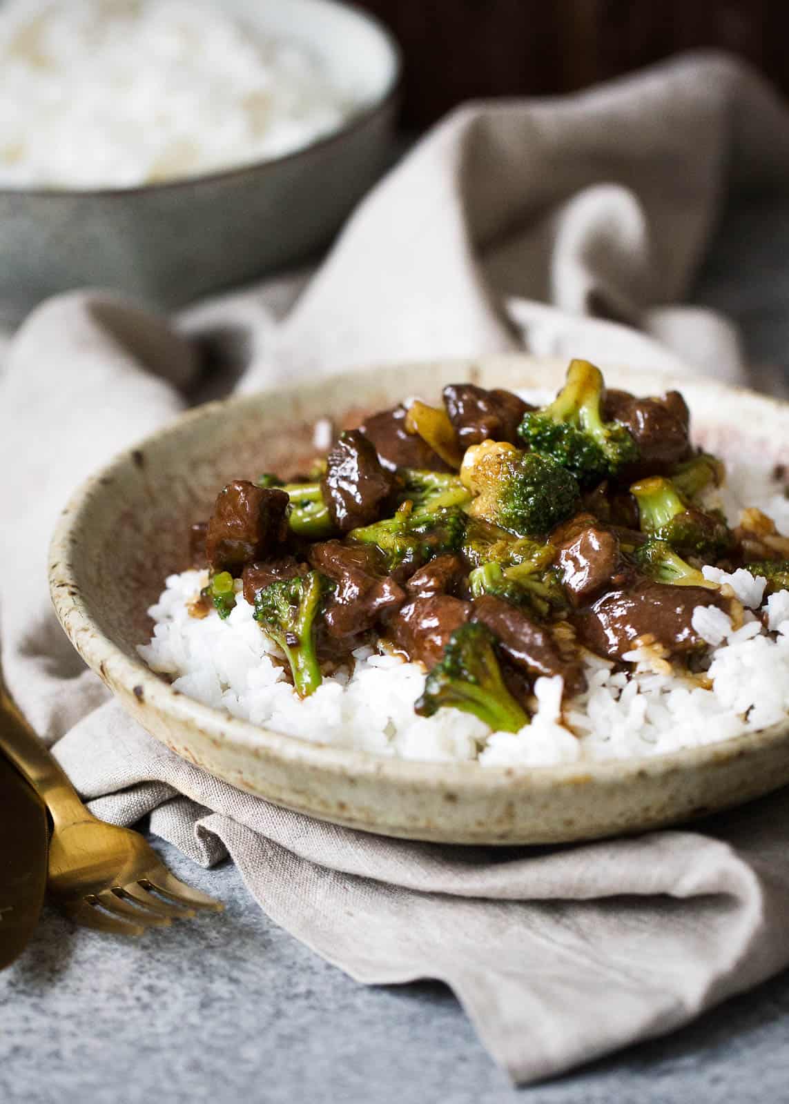 beef and broccoli mixture on top of white rice in a brown and tan speckled bowl set atop a beige linen towel. there is white rice in a dark grey bowl in the background along with a gold fork near the beef and broccoli