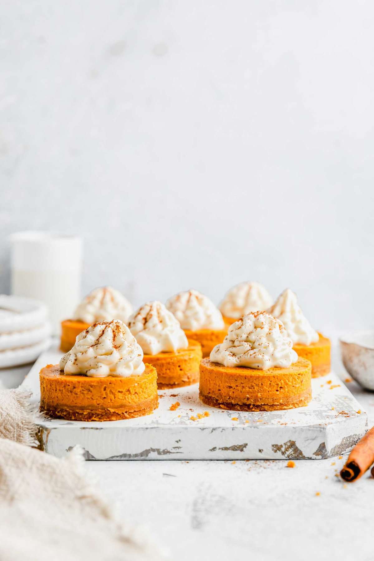 A tray filled with mini pumpkin cheesecake bites, dusted in cinnamon, with a tray and pitcher in the background, and a kitchen towel and cinnamon stick in the foreground