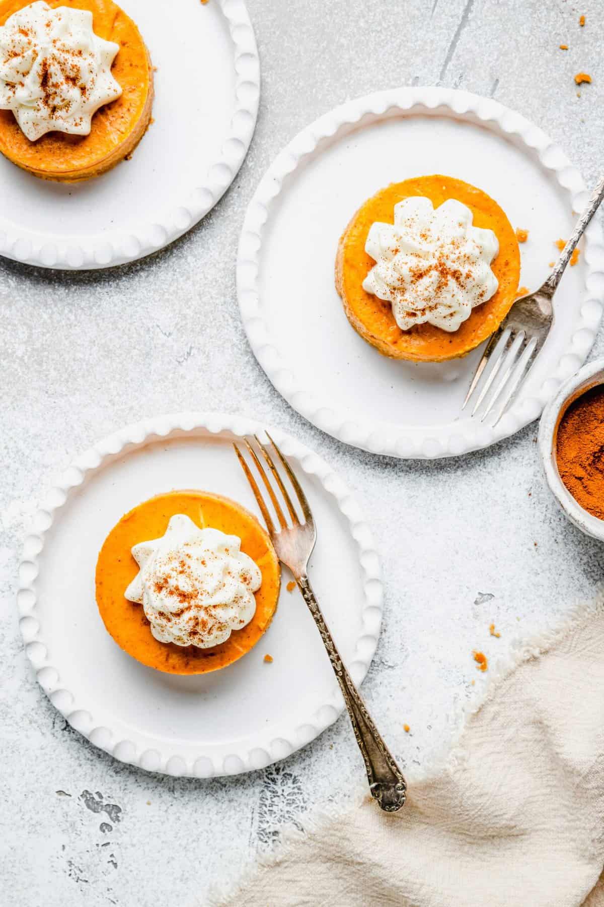 Three plates with a mini pumpkin cheesecake bite on them, dusted with cinnamon, with a fork on each plate, next to a bowl of ground cinnamon