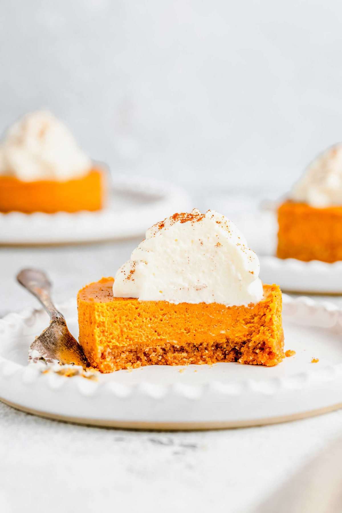 A mini pumpkin cheesecake bite on a plate, with a bite taken out of it and a fork on the plate, with two cheesecake bites on plates in the background