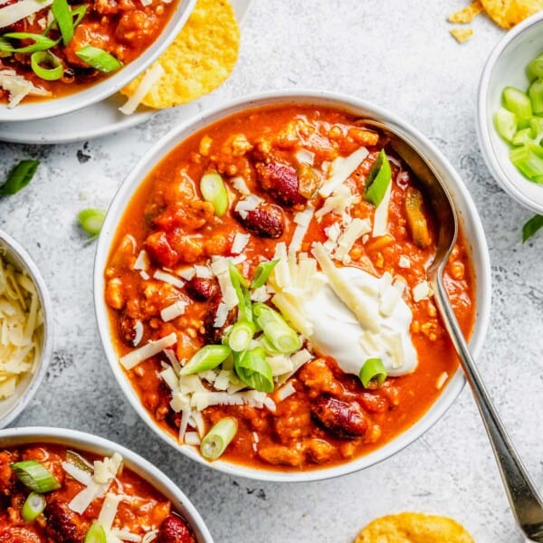 Overhead view of a bowl of turkey pumpkin chili with green onions, cheese, and sour cream on top, a spoon sticking out, and other bowls next to it, with tortilla chips all around