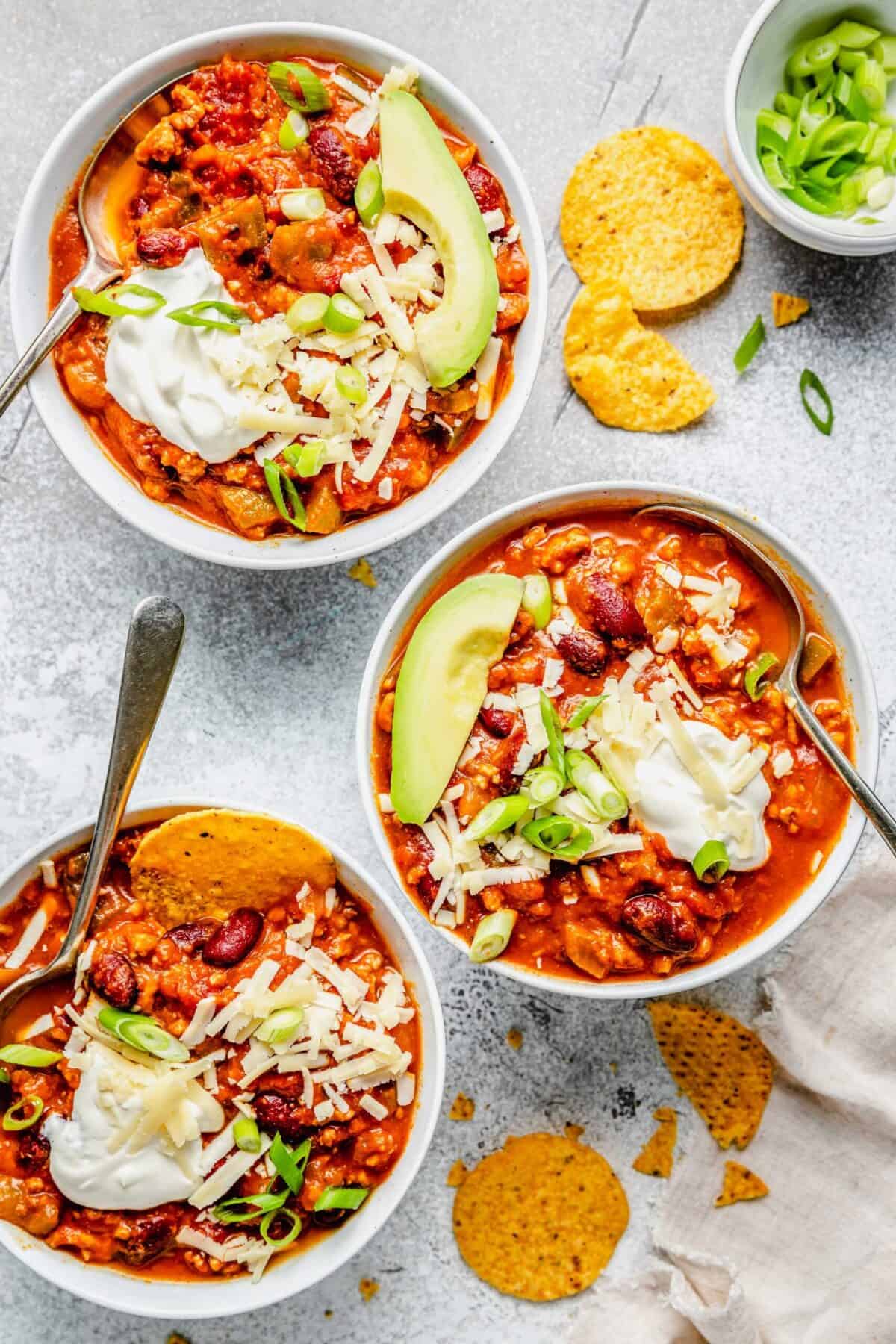 Three bowls of turkey pumpkin chili with green onions, sour cream, cheese, tortilla chips, and avocado on top, with a spoon in each bowl