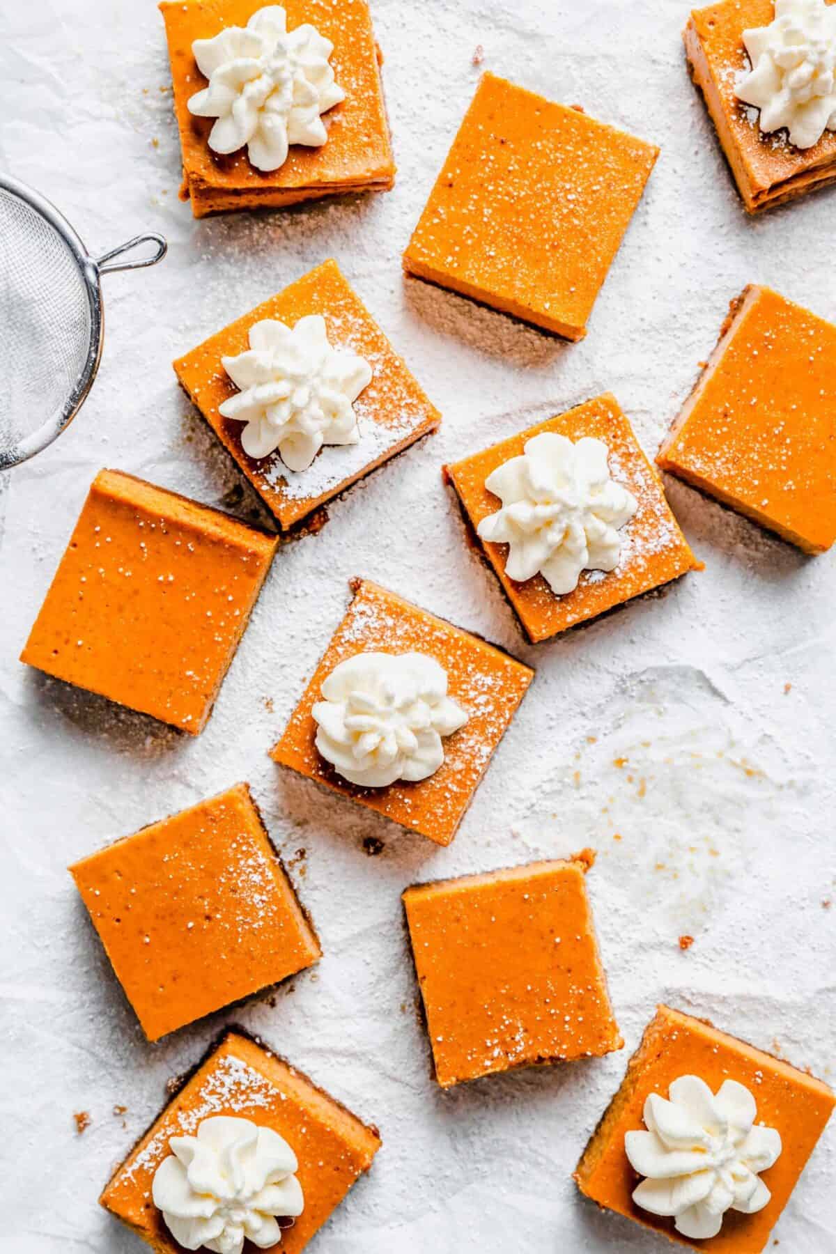12 squares of maple pumpkin cheesecake bars, some with powdered sugar on top, some with dollops of whipped cream, surrounded by a sieve and powdered sugar