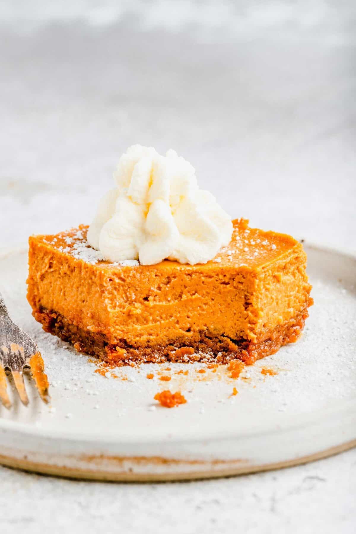 A slice of maple pumpkin cheesecake bar on a plate, topped with whipped cream, with a bite taken out of it and a fork on the plate