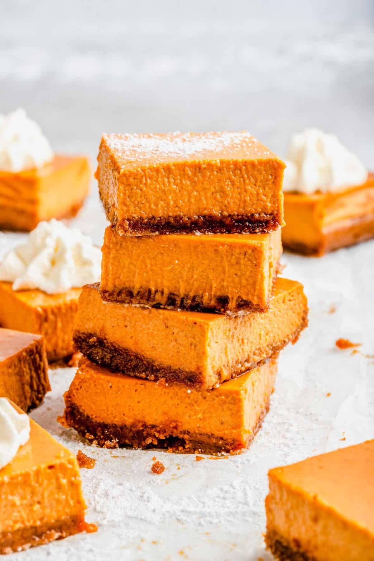A stack of four squars of maple pumpkin cheesecake bars on top of each other, dusted with powdered sugar, with whipped cream-topped bars in the background
