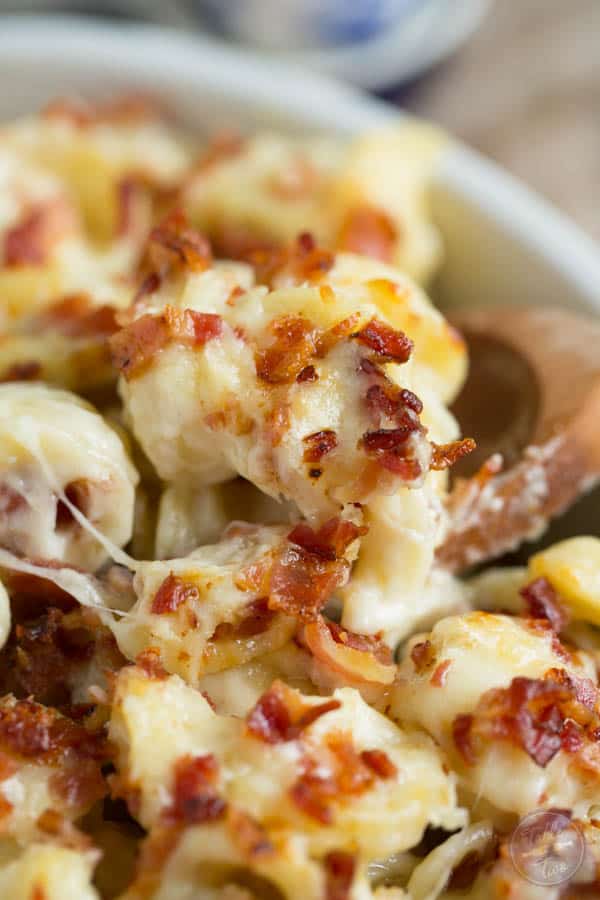 A wooden spoon scoops a serving from a mac and cheese casserole topped with crumbled bacon.