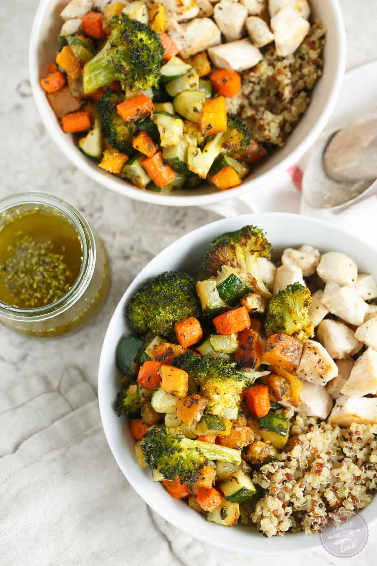 Overhead view of two quinoa bowls topped with roasted vegetables and chicken next to a jar of dressing.