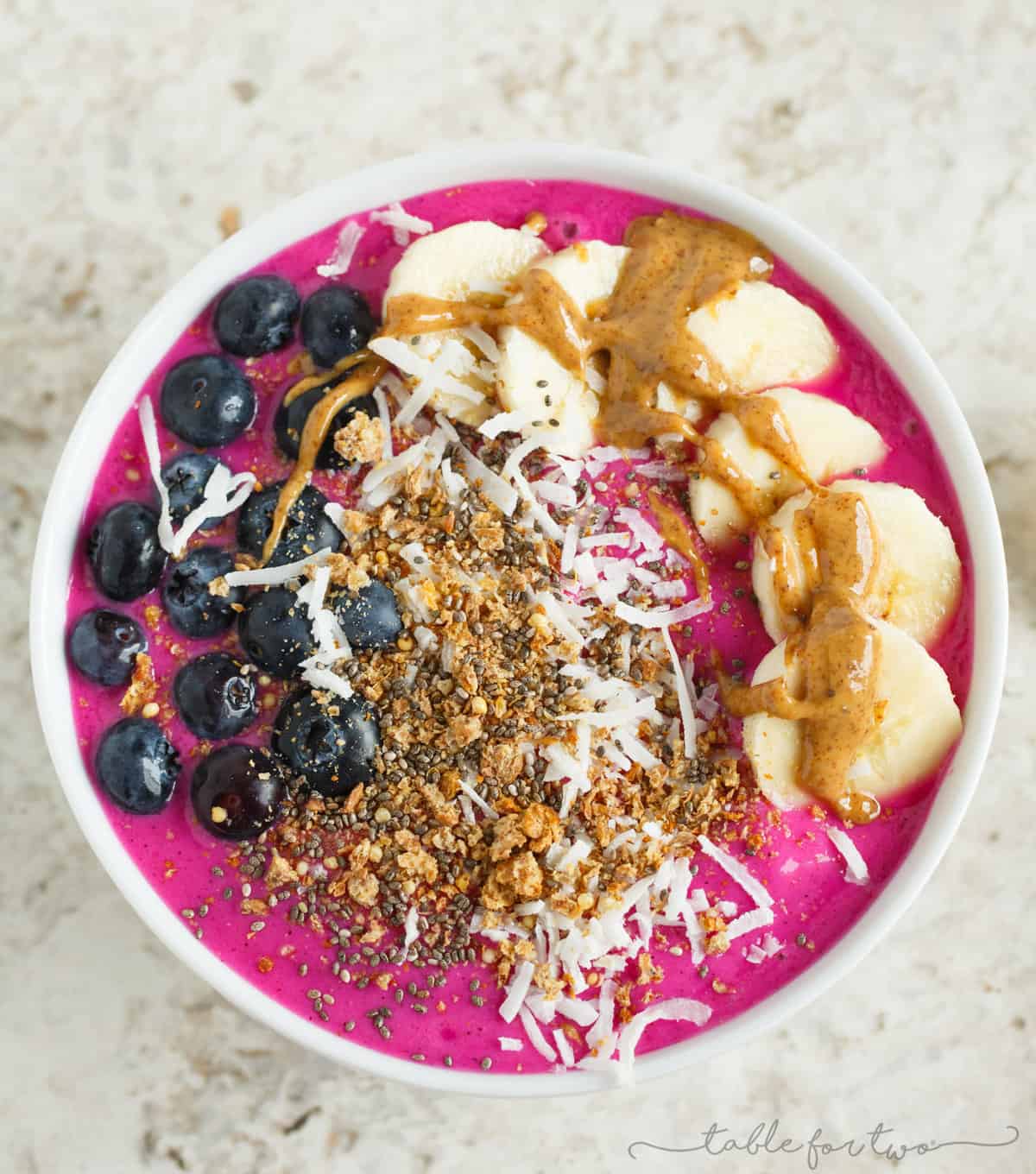 Overhead view of a pitaya smoothie bowl topped with blueberries, bananas, almond butter, coconut, and granola. 