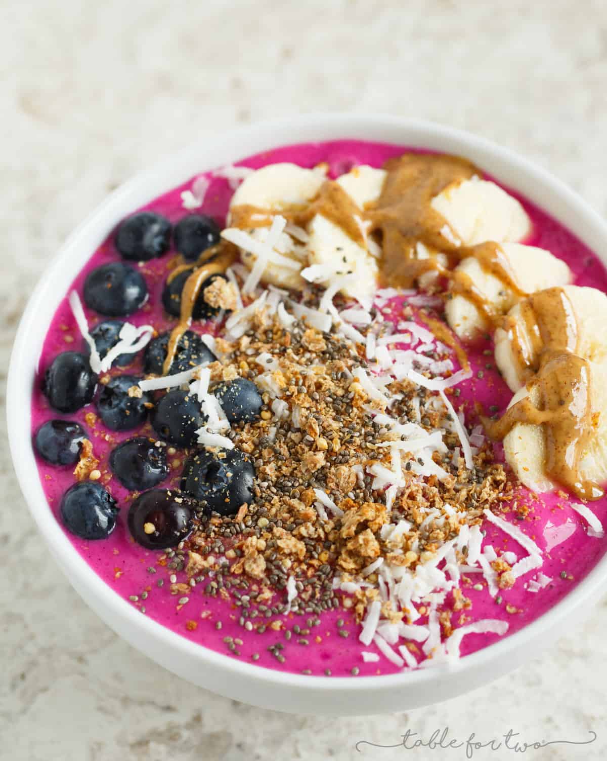 A pitaya smoothie bowl, topped with blueberries on the left side, bananas and almond butter on the right side, and granola and coconut down the middle. 