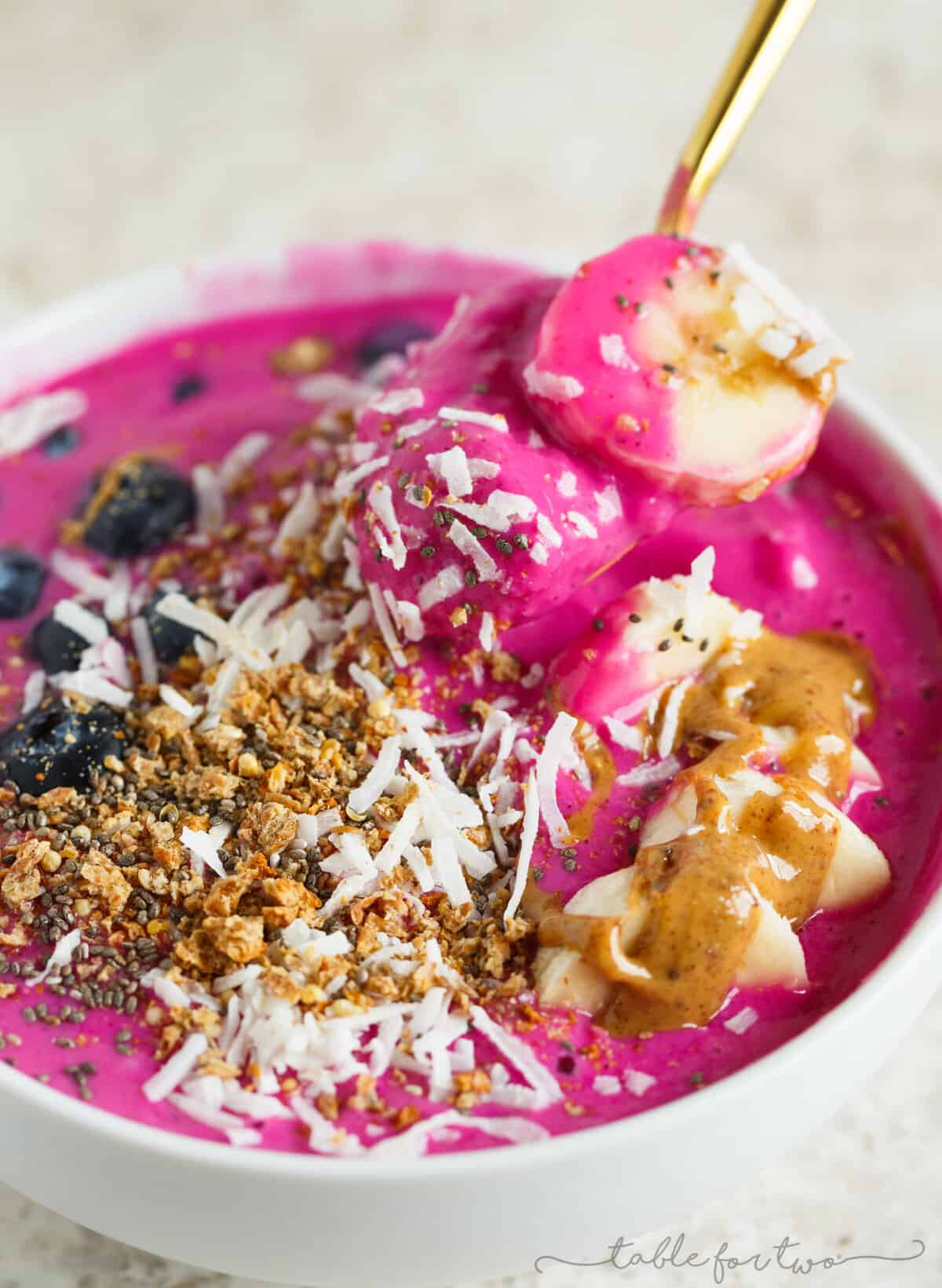 Close up of a pitaya smooth bowl topped with bananas, blueberries, granola, coconut, and almond butter, with a spoon pulling out a spoonful with bananas.