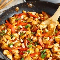 a wooden spoon is in a nonstick skillet with chicken covered in a peanut sauce surrounded by red bell peppers and green onions