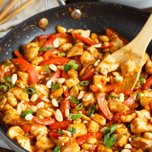 a wooden spoon is in a nonstick skillet with chicken covered in a peanut sauce surrounded by red bell peppers and green onions