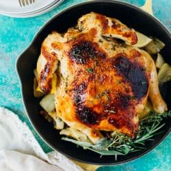 Pressure cooker honey butter and herb roasted chicken with golden-brown skin sits in a cast iron skillet, garnished with herbs and surrounded by roasted vegetables. Plates and forks rest beside the skillet on a blue surface.