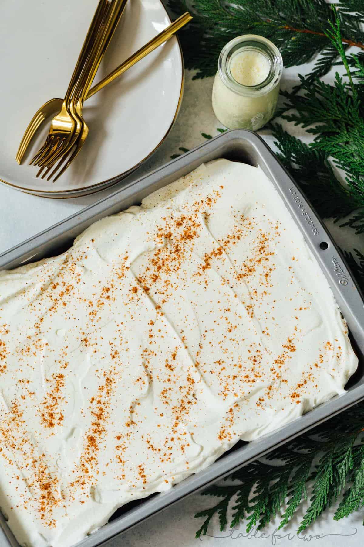 Eggnog tres leches cake in the pan next to pine leaves and plates with gold forks.