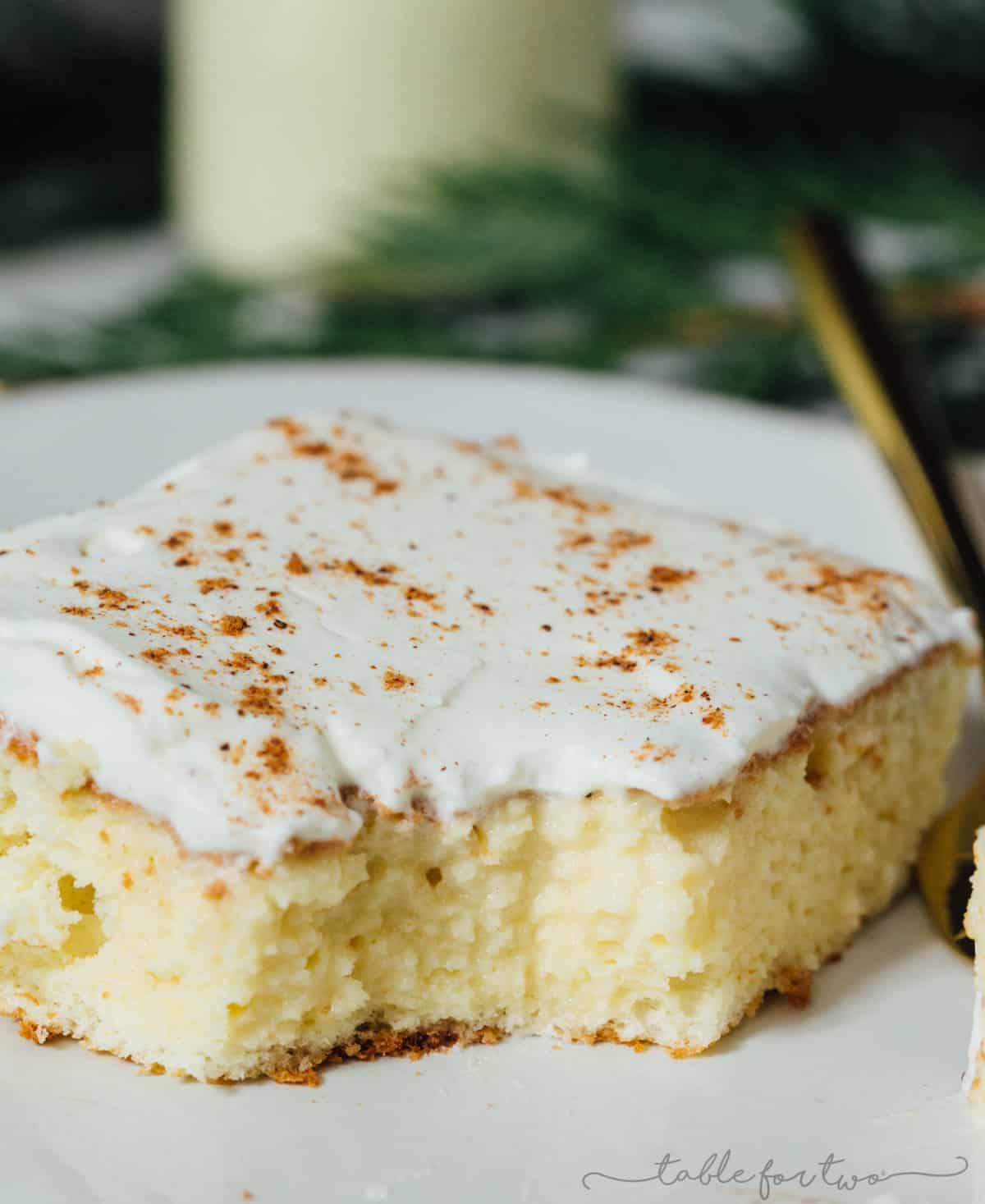 Slice of eggnog tres leches cake with whipped topping and nutmeg sprinkled on top. In the photo, the cake slice has a corner taken out to show the moist center.