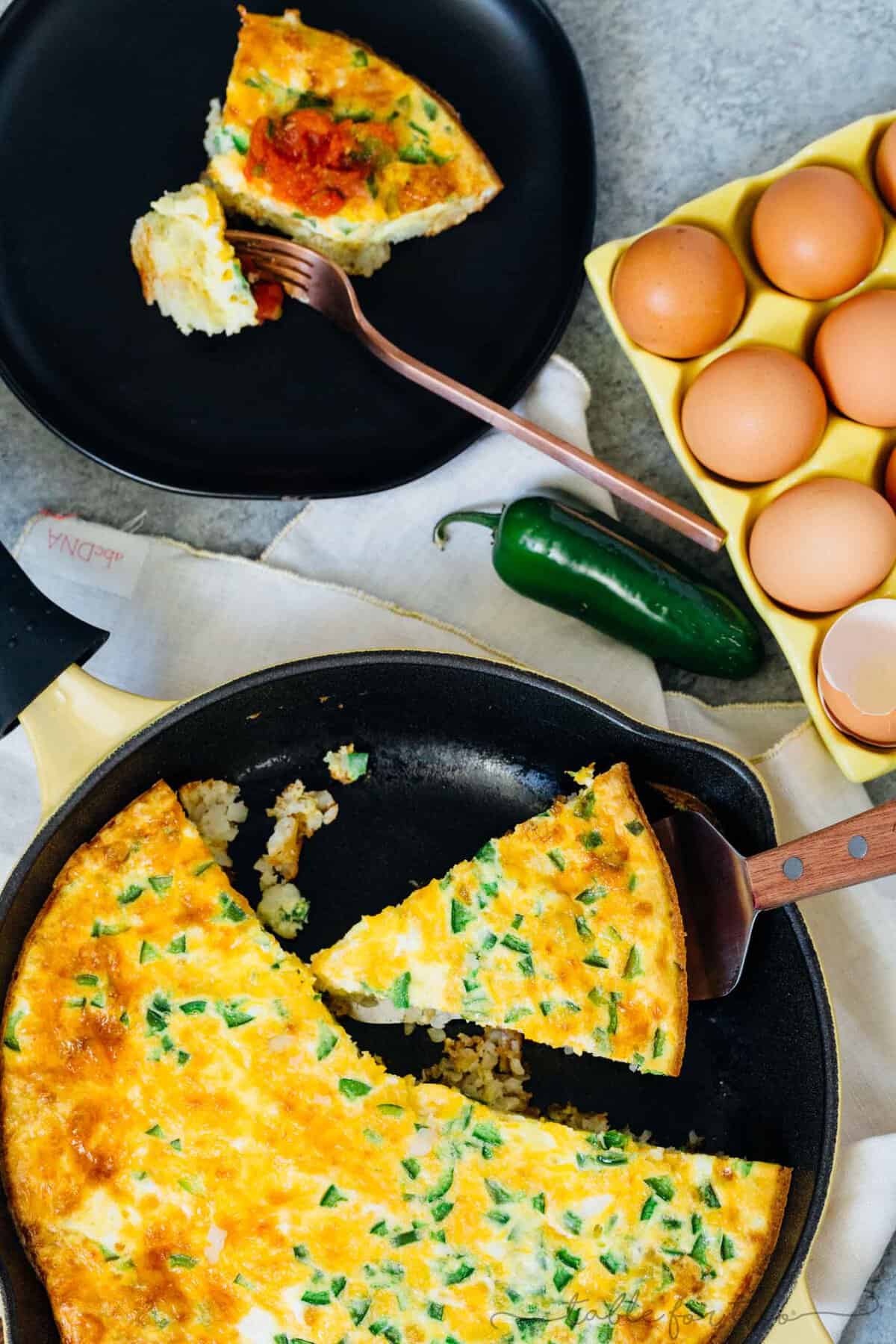 Overhead view of a slice of tater tot egg frittata next to the rest of a frittata in a cast iron skillet, next to another slice of frittata on a black plate and a carton of eggs.