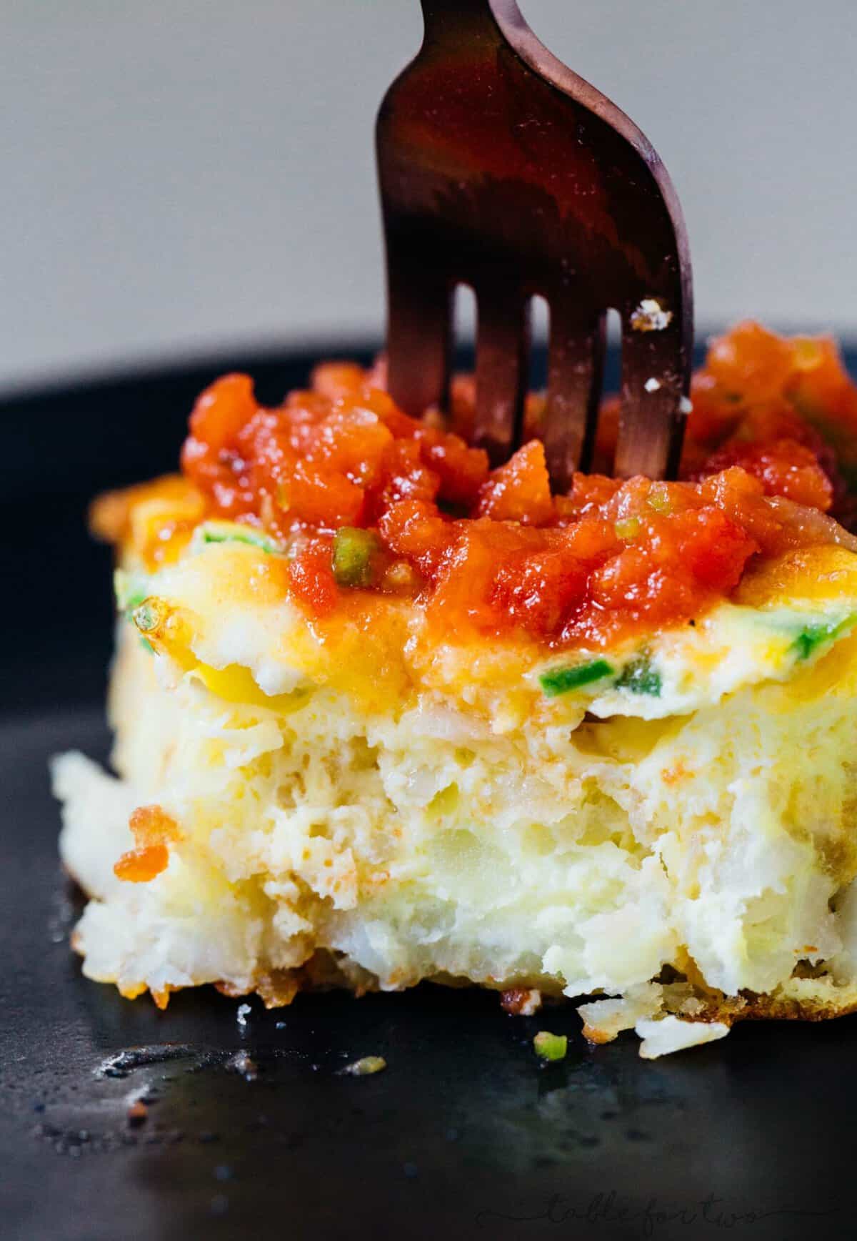 This one-pan cheddar jalapeno tater tot frittata recipe puts a fun twist on a potato and egg frittata, baked inside a crispy tater tot crust. A fork stuck into a slice of cheddar jalapeño tater tot frittata on a black plate.