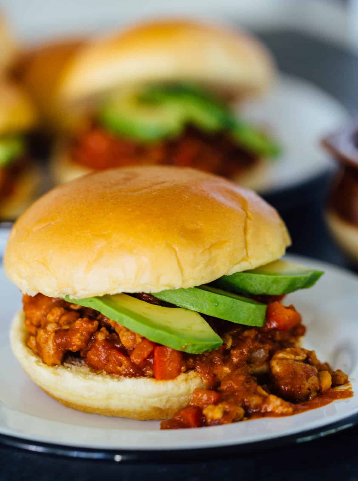 A pumpkin BBQ sloppy joes sandwich garnished with avocado slices, spilling out of the bun on a plate, with another plate full of sloppy joes in the background.