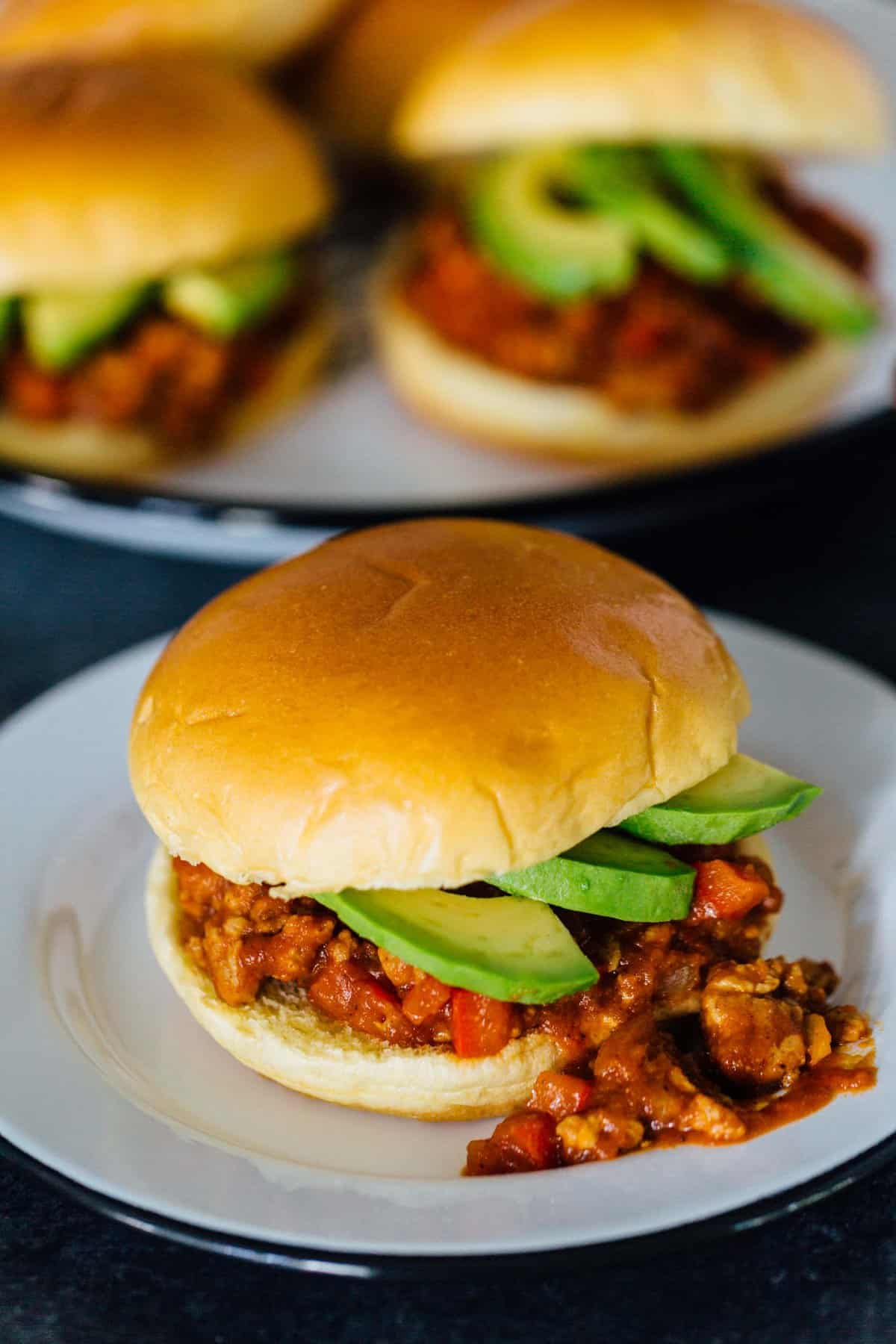 A pumpkin BBQ sloppy joes sandwich garnished with avocado slices, spilling out of the bun on a plate, with another plate full of sloppy joes in the background.