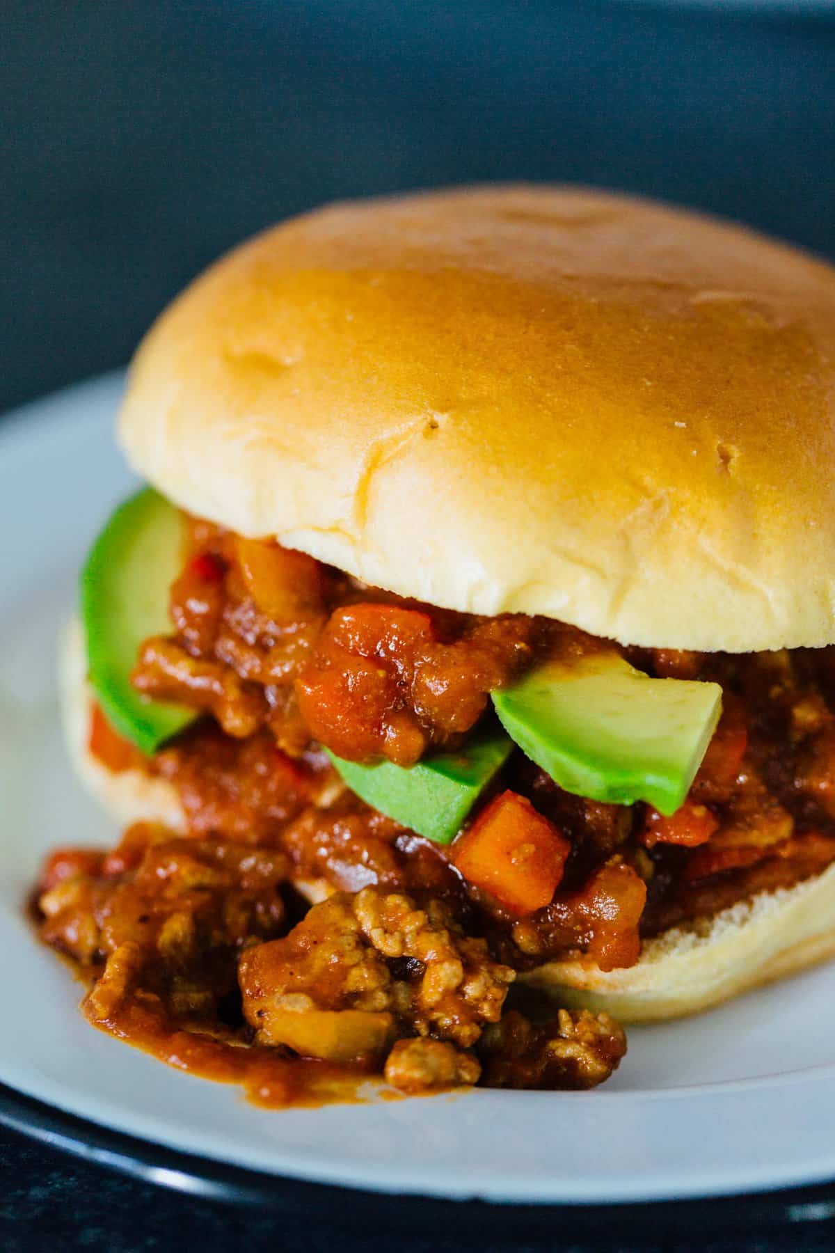 A pumpkin BBQ sloppy joes sandwich garnished with avocado slices, spilling out of the bun on a plate.