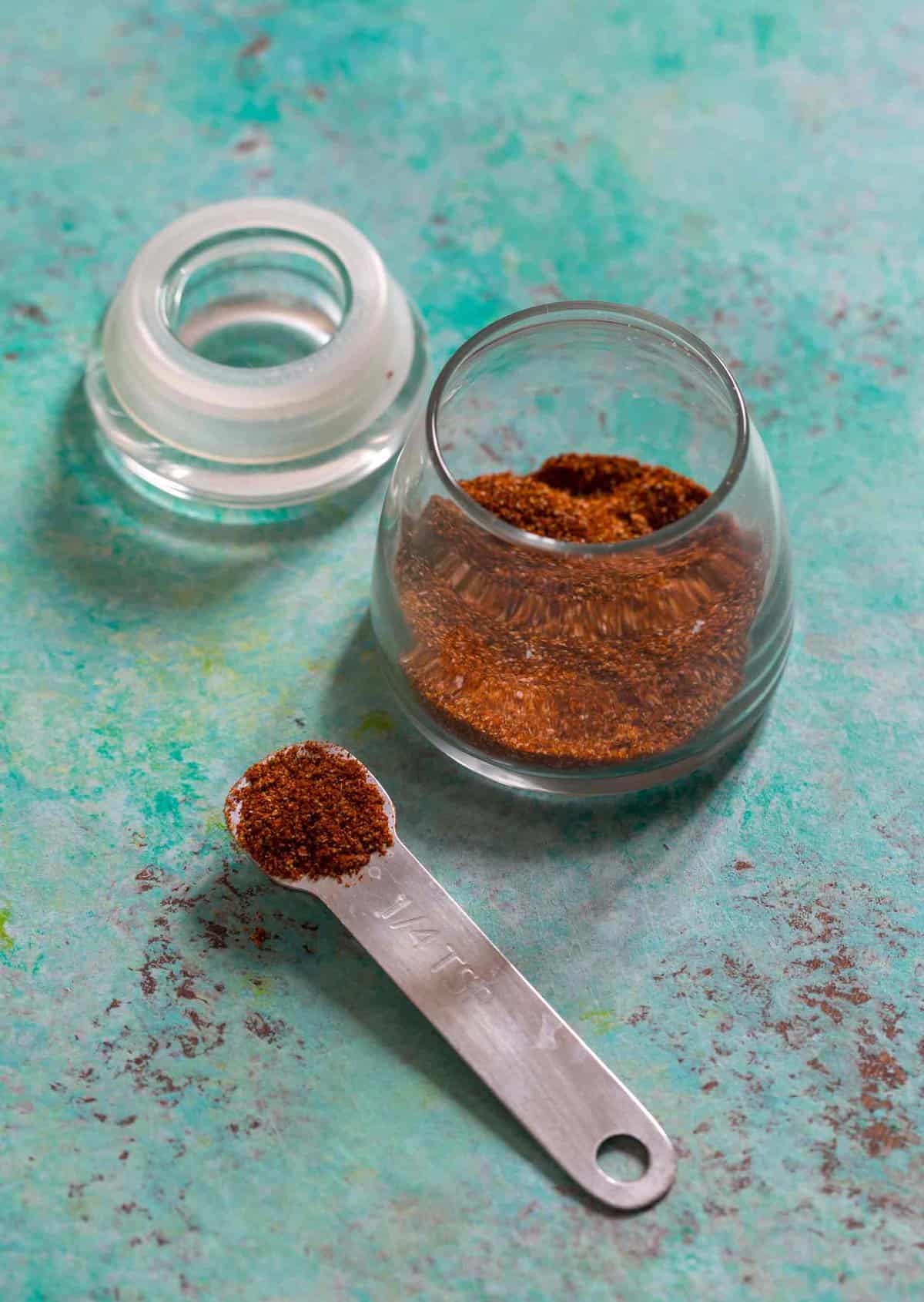 Small measuring spoon filled with fajita seasoning, with glass jar of seasoning in background