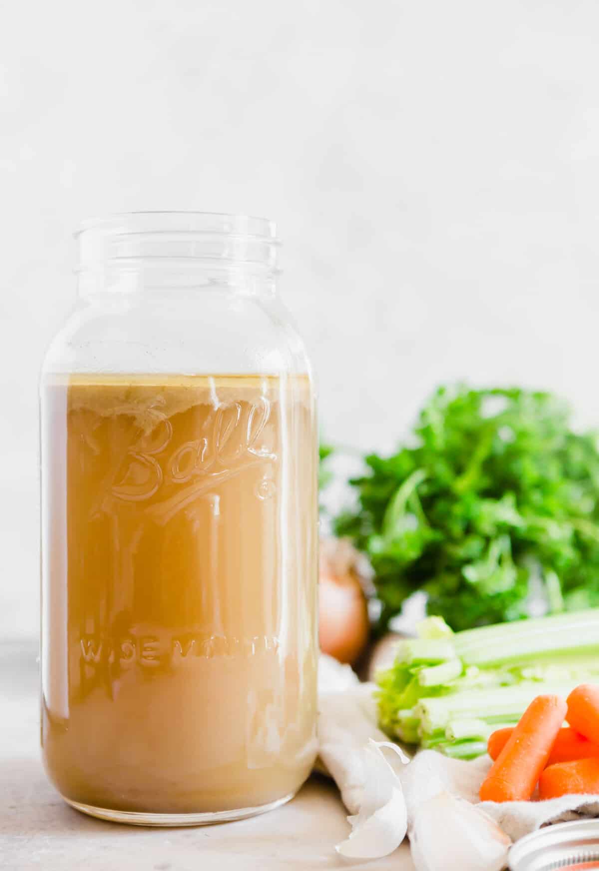 a large wide mouth Ball mason jar filled with chicken stock next to fresh ingredients like parsley, carrots, and celery.