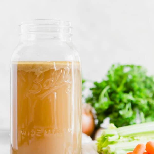 a large wide mouth Ball mason jar filled with chicken stock next to fresh ingredients like parsley, carrots, and celery.