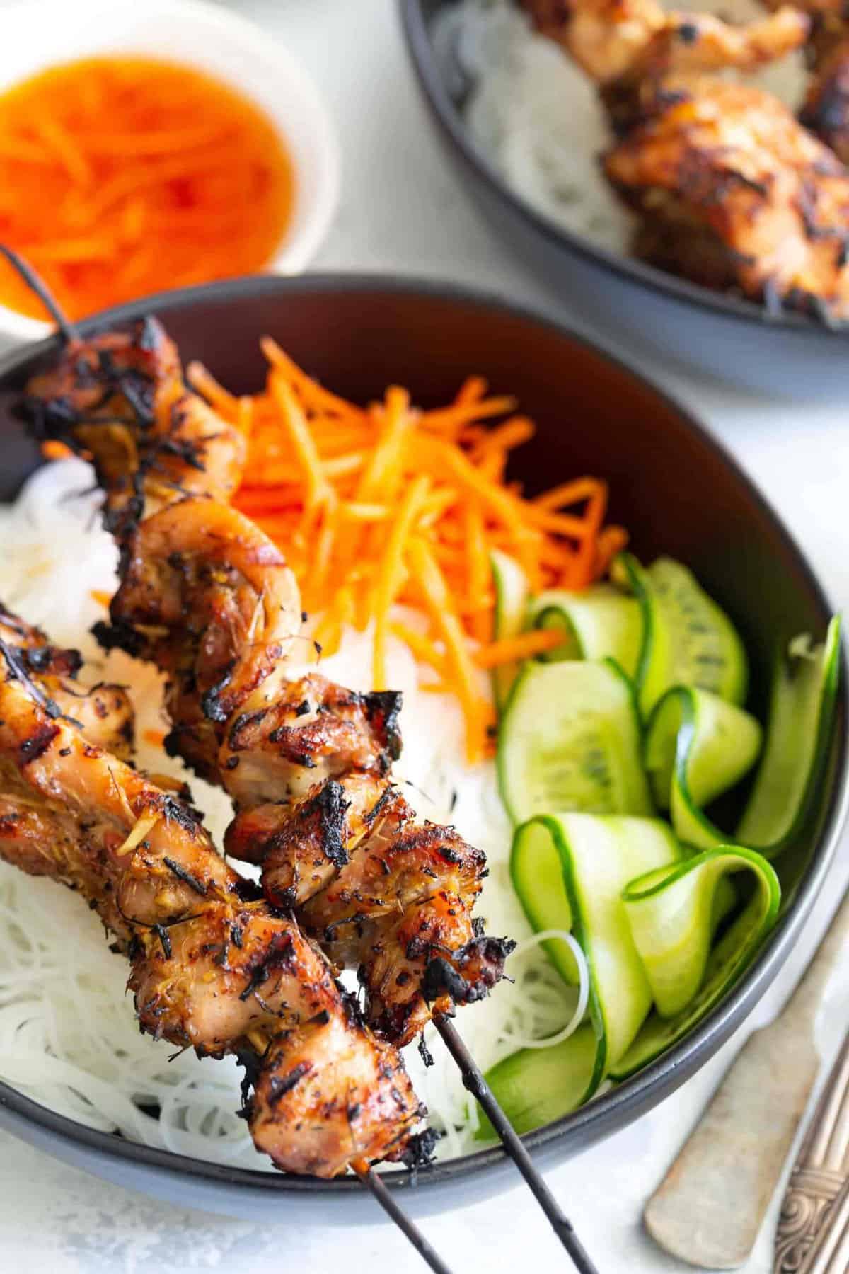 Grilled lemongrass chicken skewers on top of rice noodle bowl with matchstick carrots and shaved cucumber