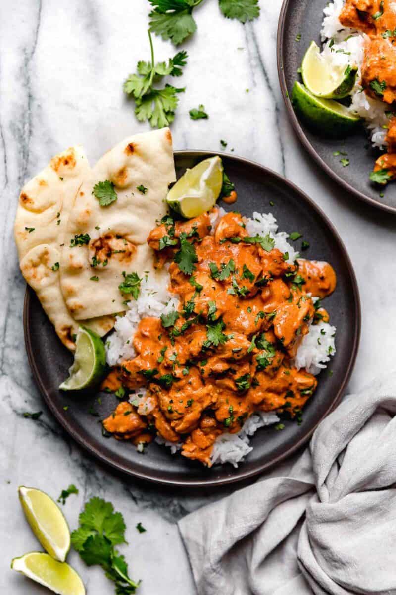 overhead image of slow cooker chicken tikka masala on a bed of white rice and a side of naan all on a dark brown plate. there is another plate of chicken tikka masala in the upper right corner.