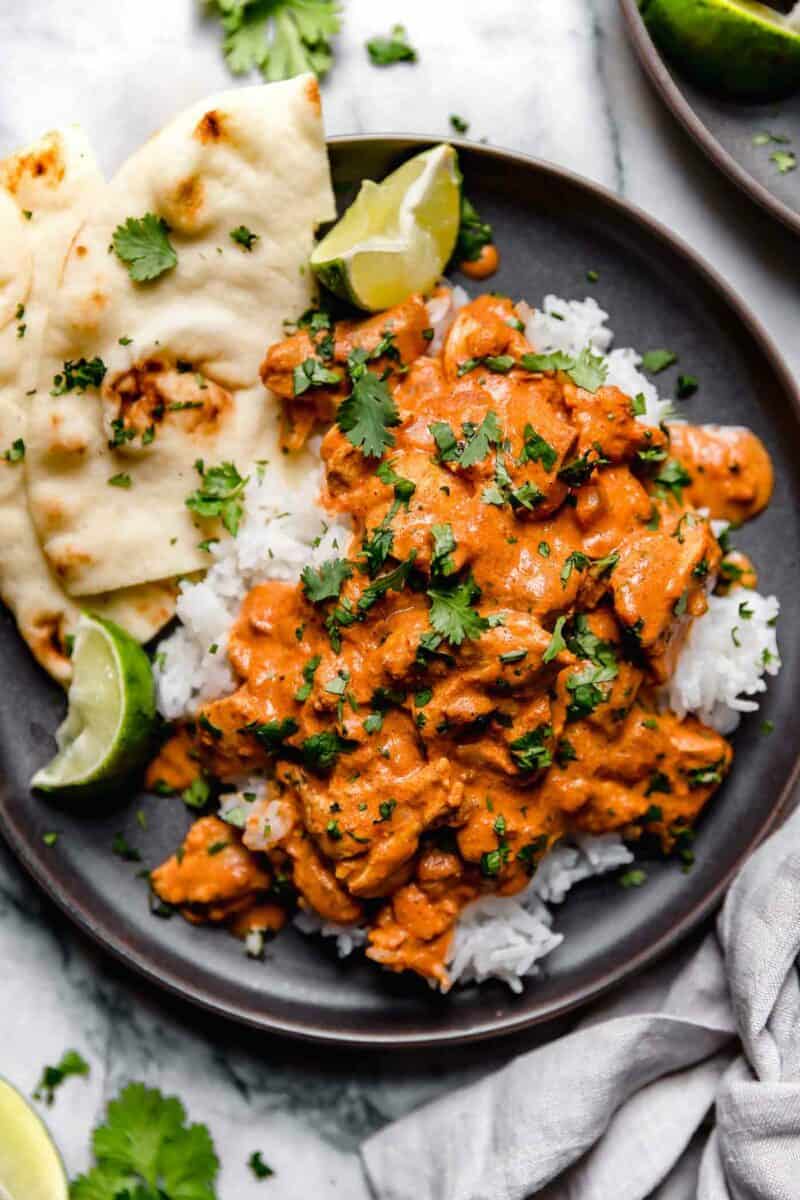 Overhead image of slow cooker chicken tikka masala on a dark brown plate on top of a bed of rice with naan on the side. The dish is garnished with lots of fresh parsley.