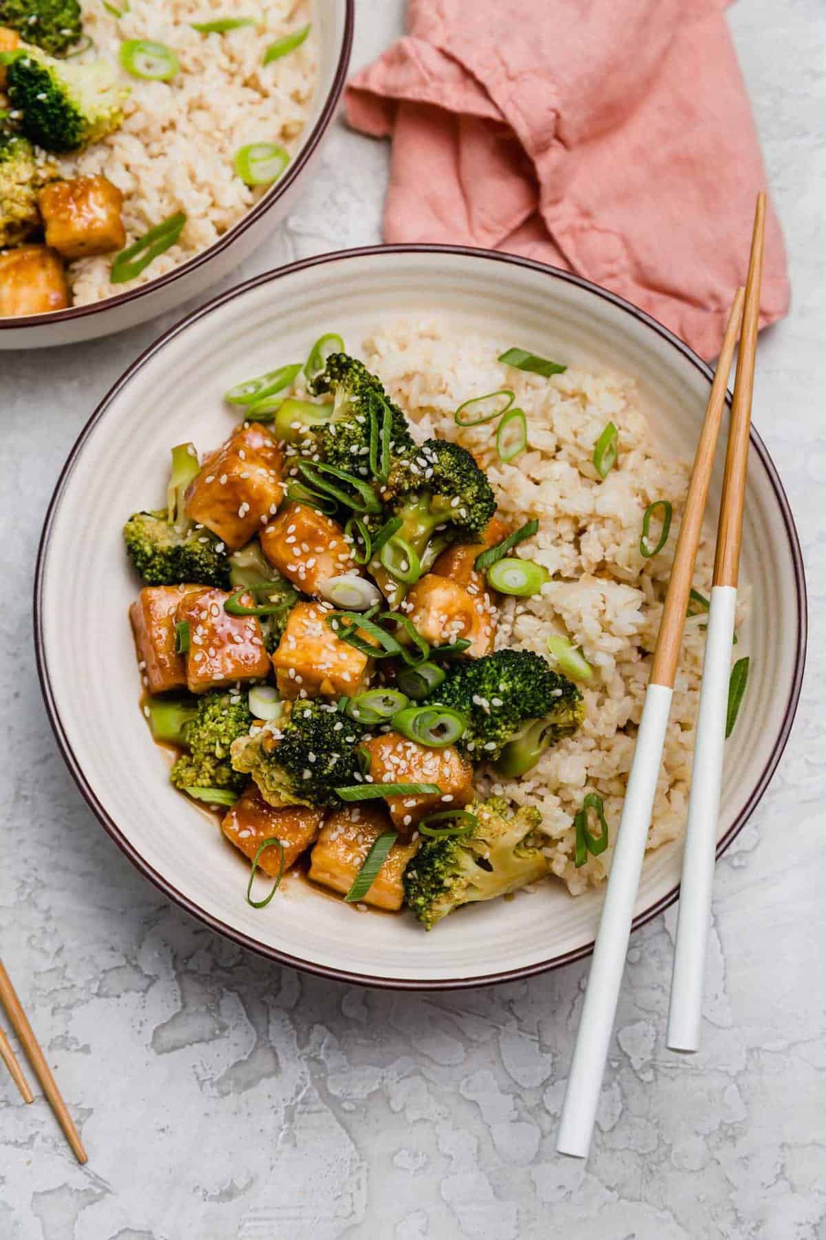 Overhead view of a teriyaki tofu bowl with broccoli, with a pair of chopsticks resting on the edge of the bowl.