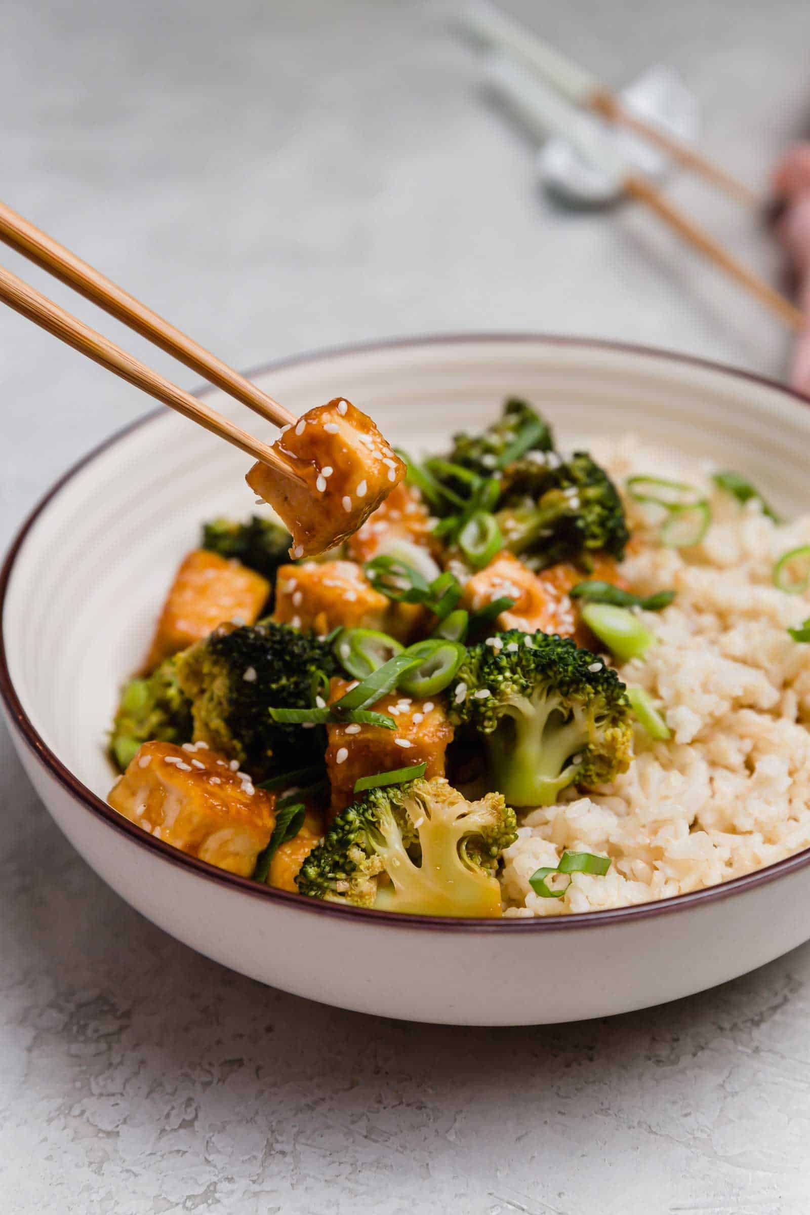 Chopsticks lifting a piece of teriyaki tofu from a bowl of tofu, broccoli, and rice.