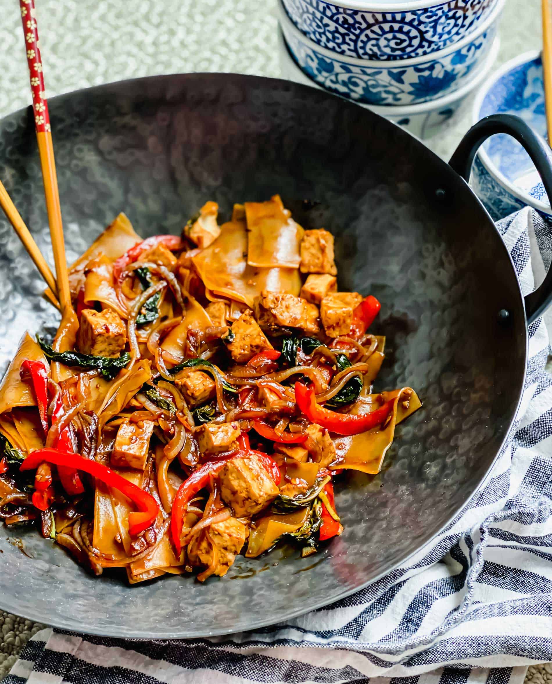 Drunken noodles in wok with with chopsticks and bowls in background.