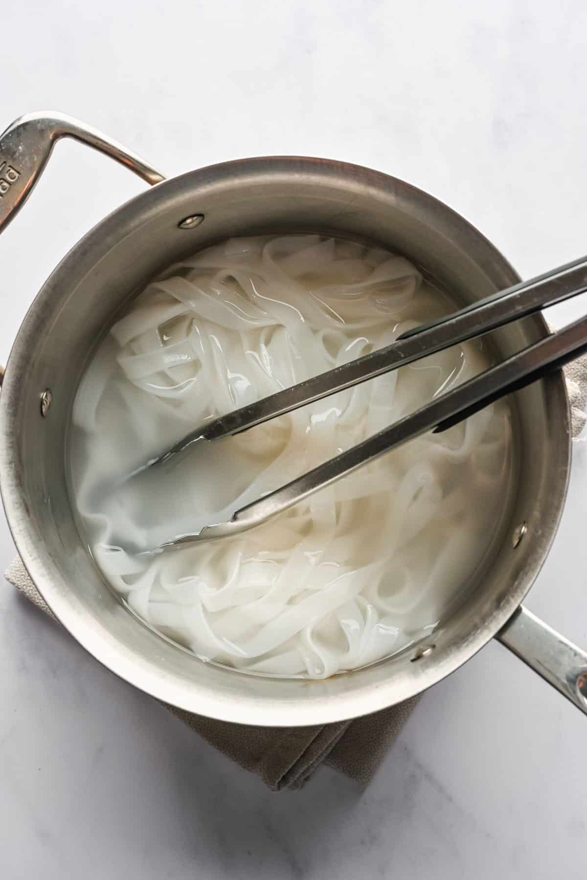 A pair of tongs resting in a pot of cooked rice noodles.