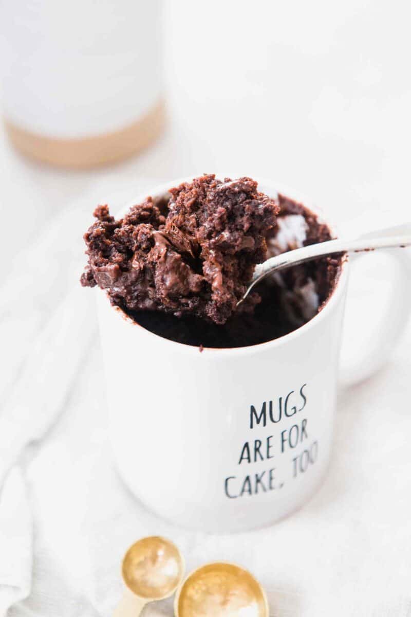 The moistest chocolate mug cake you will ever have! It's not spongy like other mug cakes! Recipe on tablefortwoblog.com A large spoonful of microwaved chocolate mug cake sitting balancing on top of a white mug that says "Mugs Are For Cakes, Too" and gold measuring spoons at the bottom of the image.