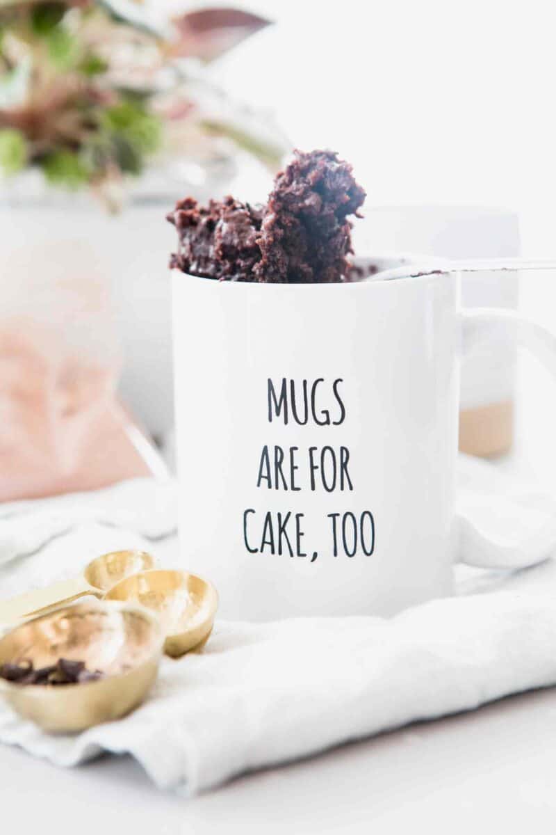 A spoonful of chocolate mug cake peeking out of a white mug that says "Mugs Are For Cake, Too" on top of a white linen towel with a measuring spoon of mini chocolate chips.