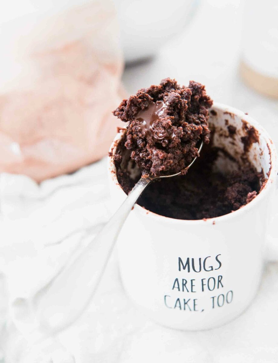 A large spoonful of chocolate mug cake sitting on top of a white mug that says "Mugs Are For Cake, Too"