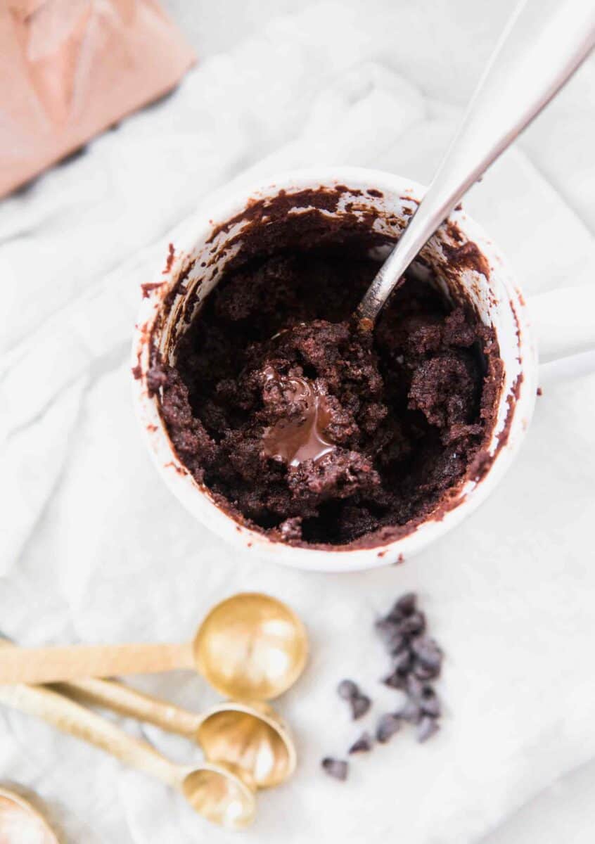 An overhead shot of the chocolate mug cake with hazelnut spread in the center. The mini chocolate chips and gold measuring spoons are on the side of the mug cake and sitting on a white linen towel.