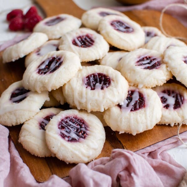 a stack of raspberry thumbprint cookies is seen on a wooden cutting board next to a pink linen towel