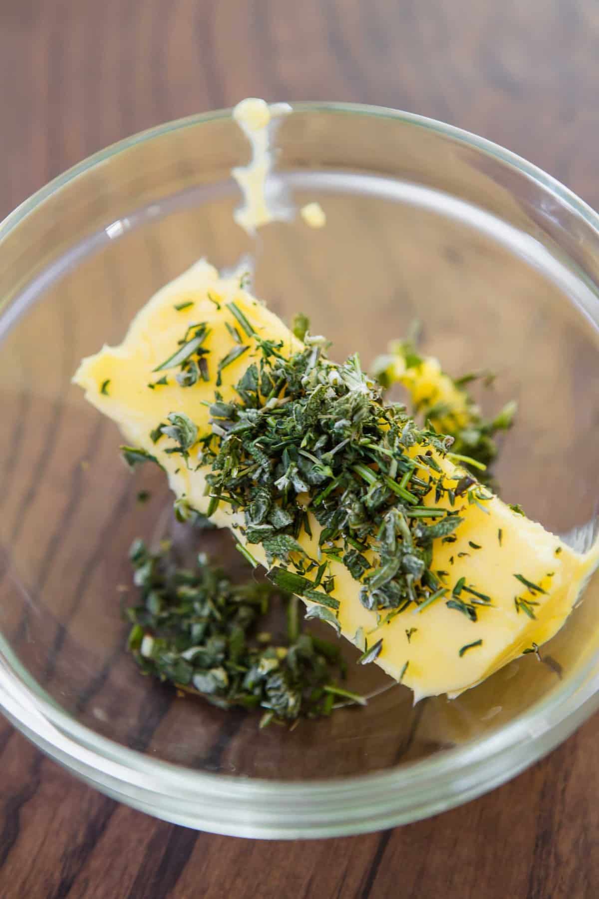 Stick of softened butter topped with coarsely chopped herbs in glass bowl