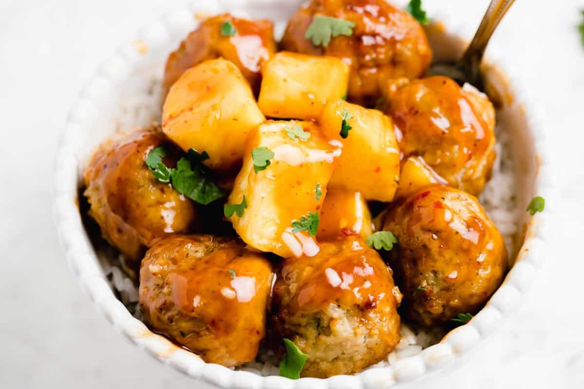 A close up shot shows sweet and sour sauce covering chunks of pineapple and meatballs in a medium sized white bowl. 
