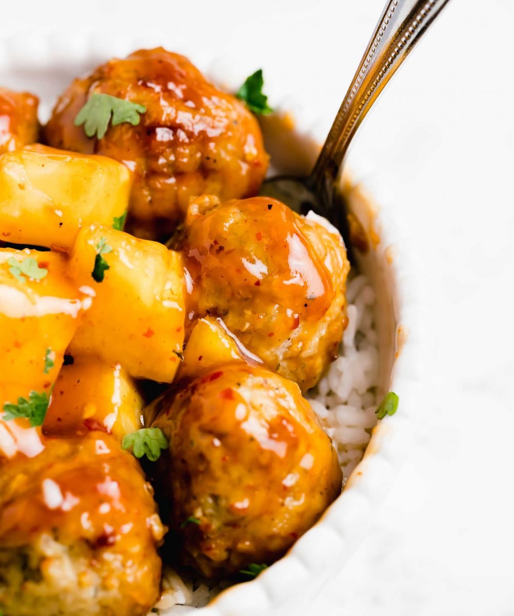 Orange sweet and sour sauce covers pineapple and meatballs in a white bowl.