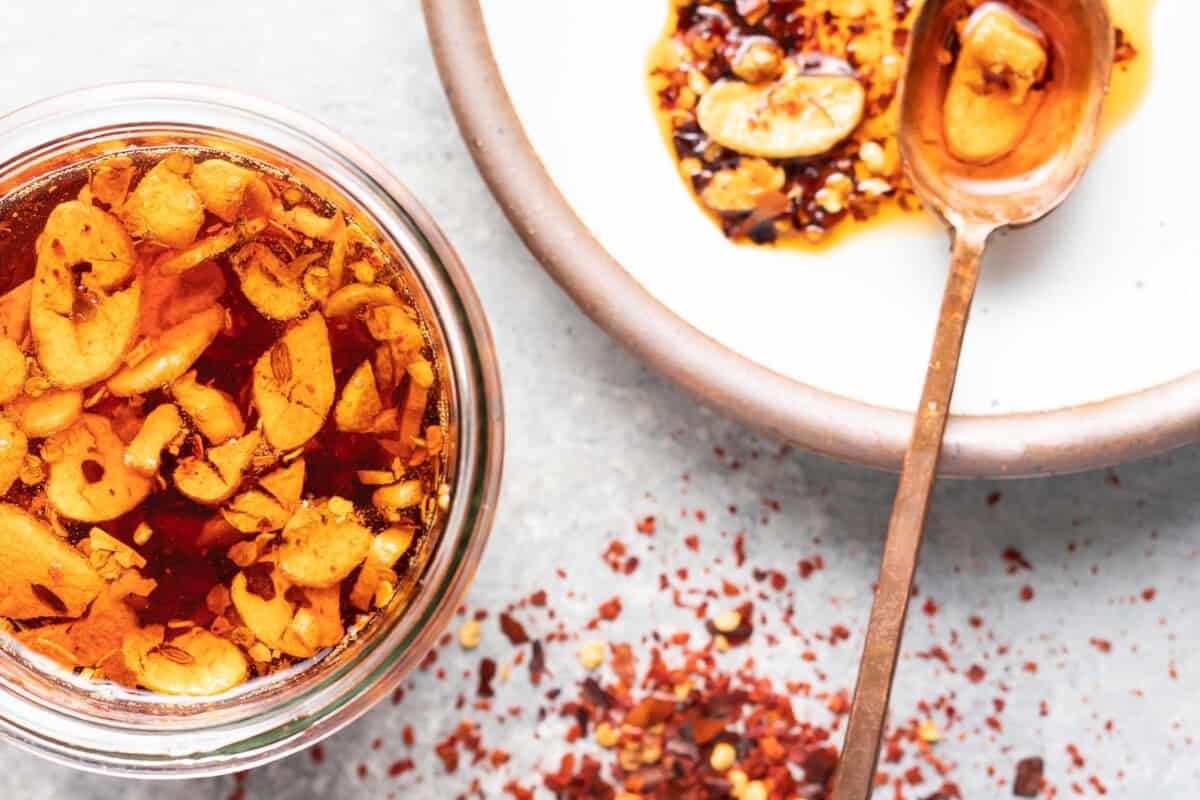 Red pepper flakes are dashed on a white surface, next to a white bowl and glass jar, both containing chili garlic oil. 