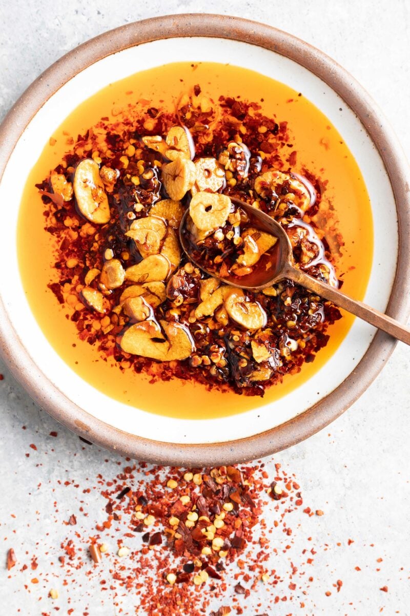 A spoon is holding a small portion of chili garlic oil above a full white bowl.