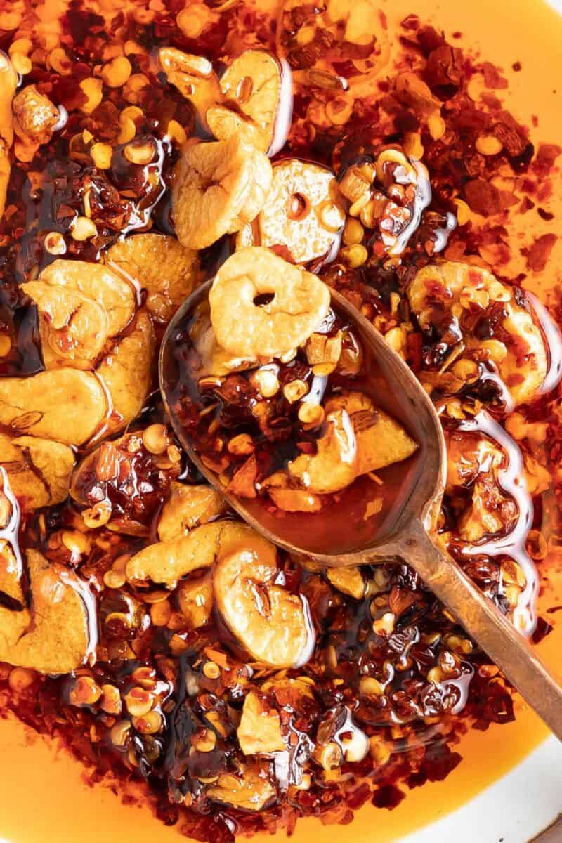 A close up image shows red pepper flakes, sliced garlic and oil in a wooden spoon.