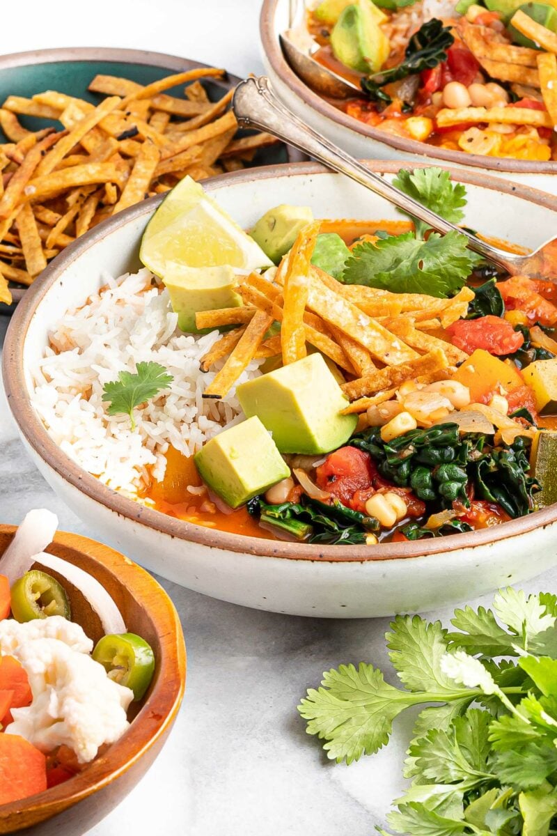 Cilantro and tortilla strips have been placed next to a full bowl of Mexican soup on a white surface. 