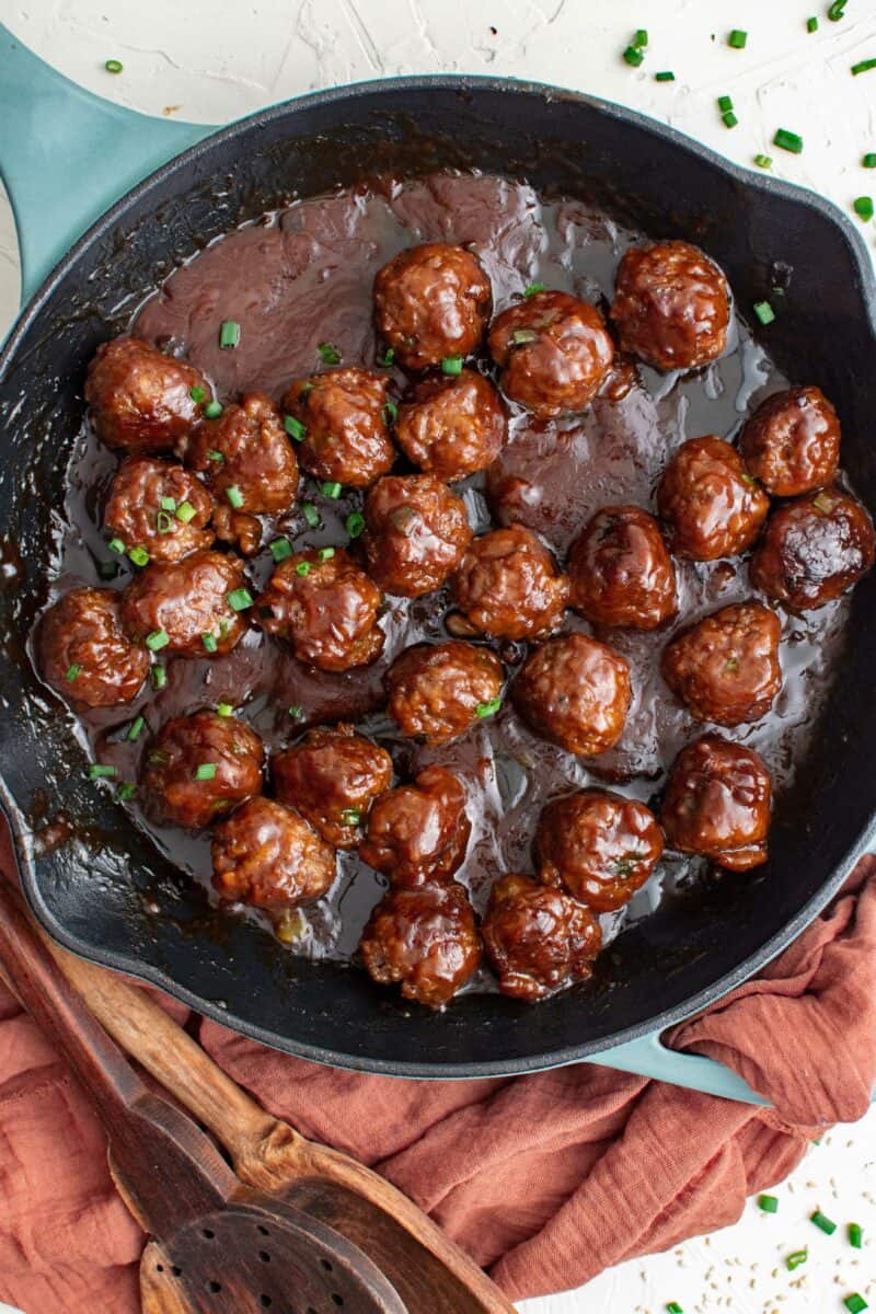 A large black skillet contains multiple cooked meatballs in a sticky dark brown sauce. 