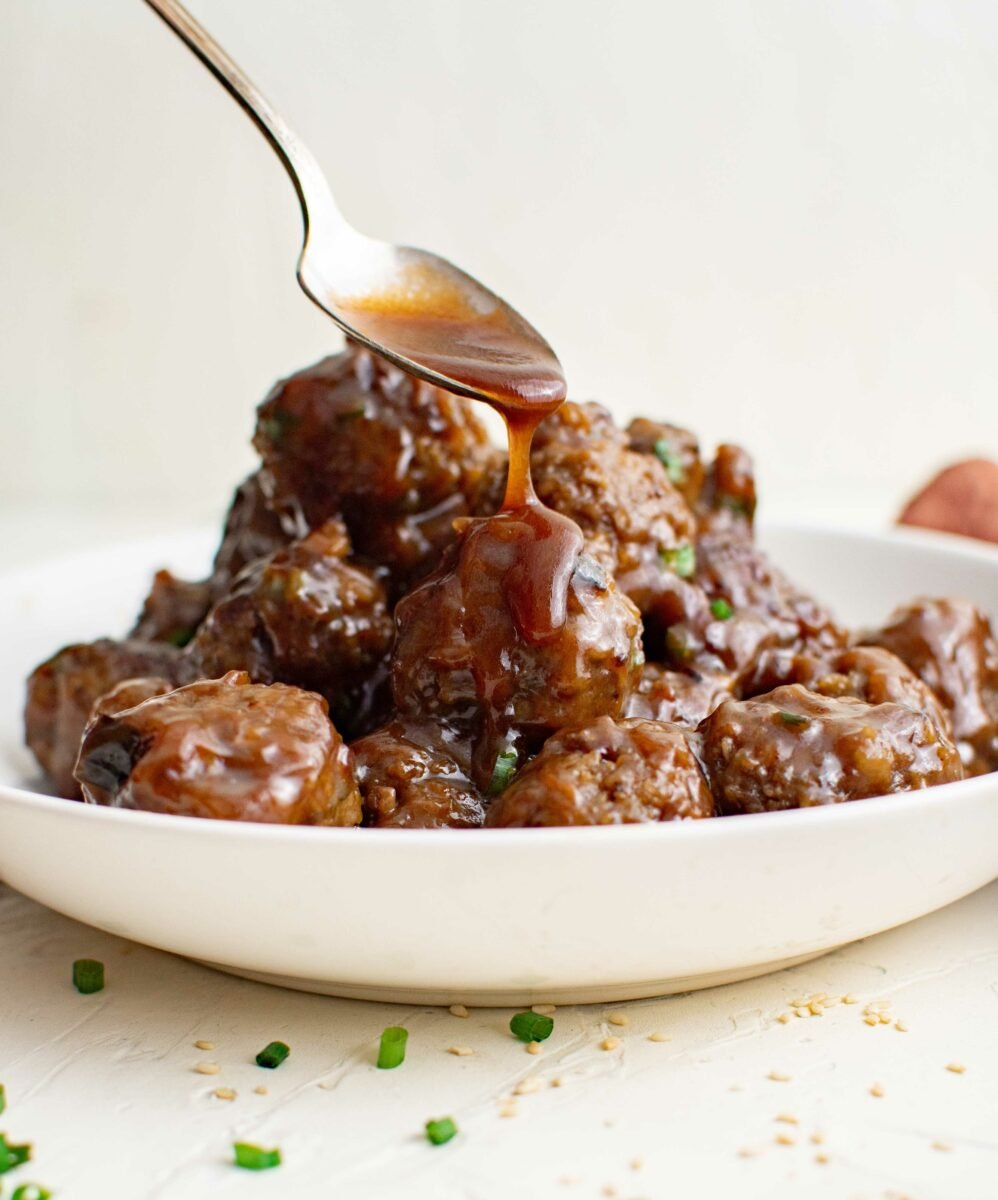 Golden brown sauce is being drizzled from a small spoon onto a pile of cooked meatballs. 